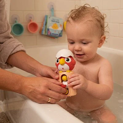 Bébé souriant dans le bain tenant un jouet hibou coloré rouge et blanc, accompagné d'un adulte — jouets de bain sécurisés pour bébé